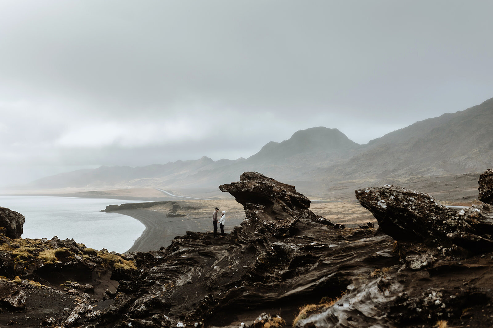 Dramatic photo of a couple climbing up on a slippery stone in a cave that has a waterfall and rainbow in Iceland