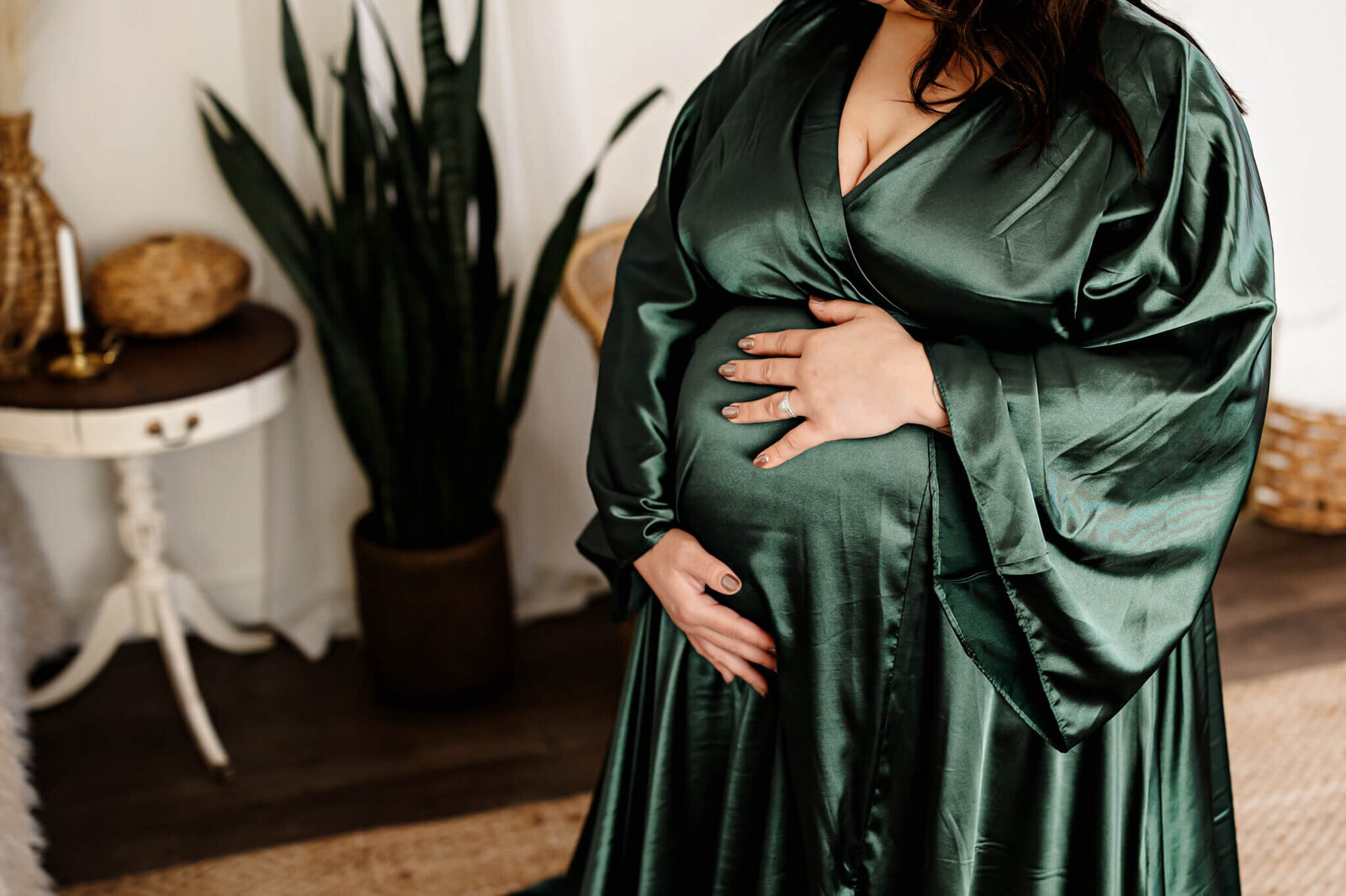 A woman in a dark green satin robe cradles her pregnant belly with both hands, standing near a potted plant and wicker baskets—an intimate moment captured by a Bloomington maternity photographer. Her face is not visible.