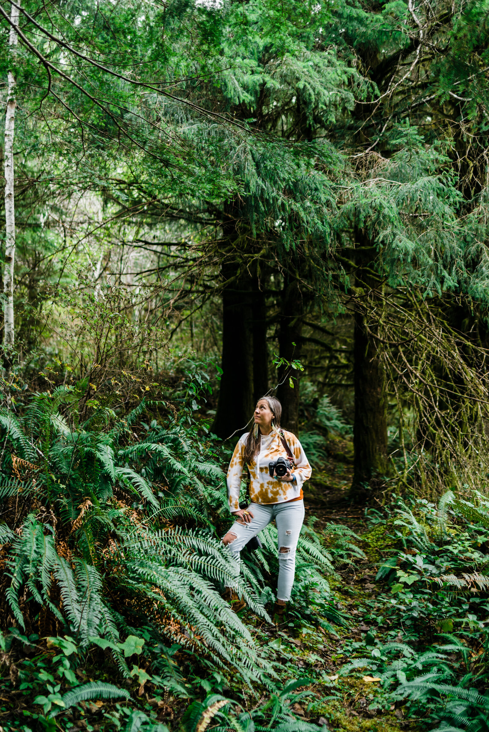 A woman with long brown hair wearing an orange tie dyed crop sweatshirt and light blue jeans with fringe moccasins is holding her Canon camera in one hand while looking up at the tall tree while standing surrounded in the lush fern forest of the PNW. | Erica Swantek Photography