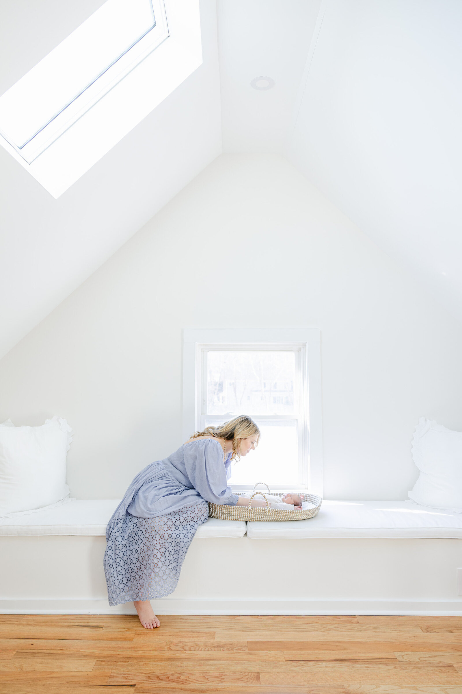 Mother in a soft blue dress leans over a newborn in a Moses basket during a peaceful lifestyle newborn session in a light-filled attic nook. Captured by Nicole Neira Photography, this Greenwich, Connecticut in-home newborn shoot highlights gentle motherhood moments in a serene, minimalist space.