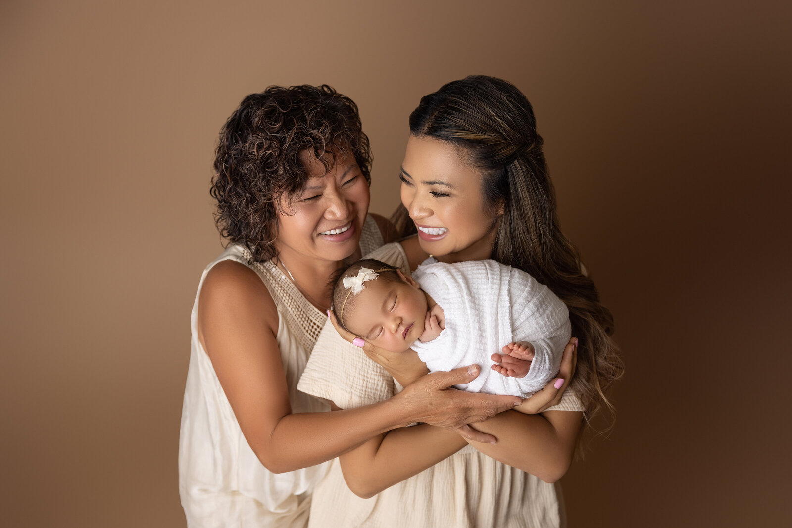 Newborn photoshoot by top London, Ontario baby photographer Amy Perrin-Ogg. Mom is holding baby in her arms with baby facing the camera. Mom is looking over her shoulder and smiling at the baby's grandma. The Grandma is smiling at the baby girl. Three generations photo.