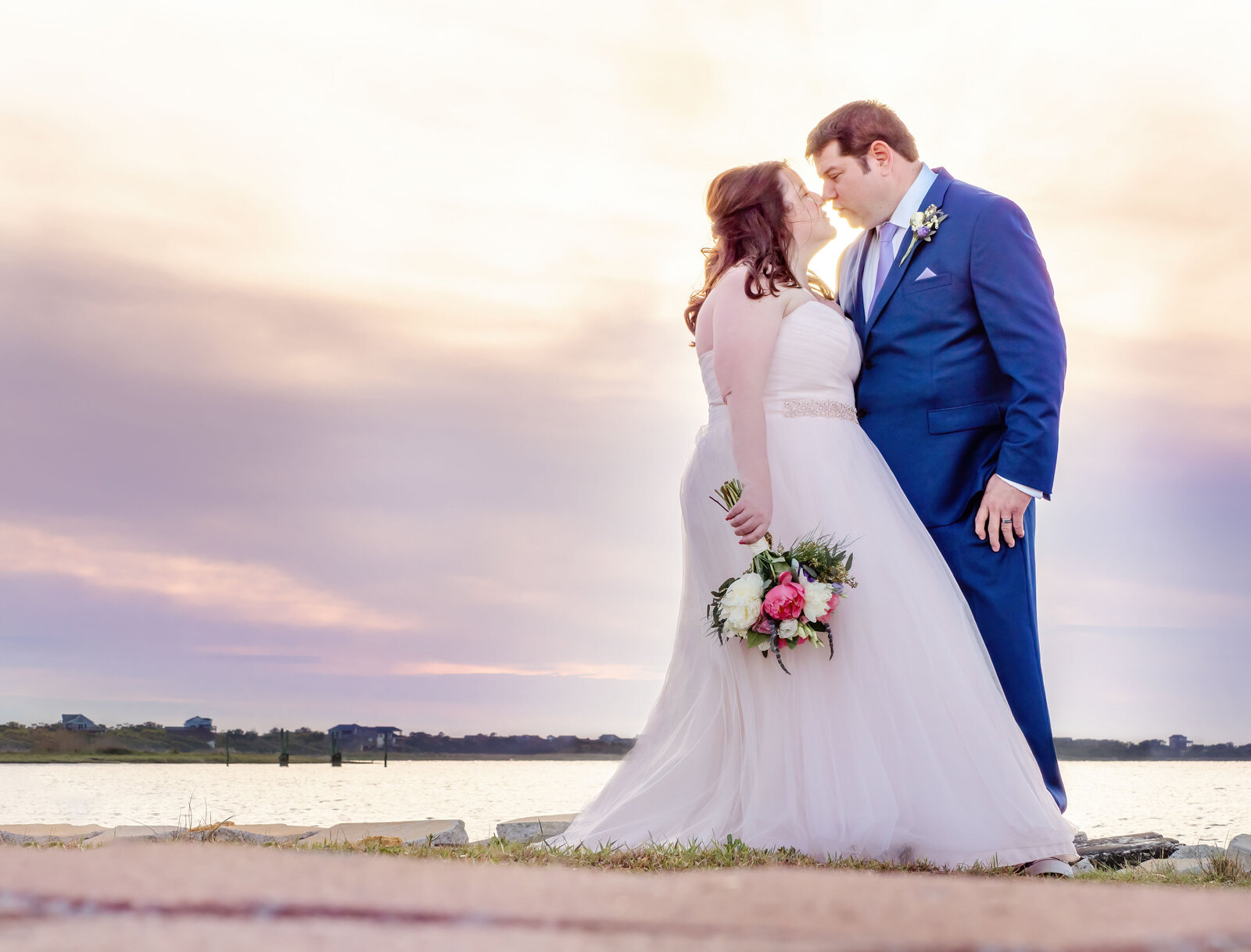 The bride and groom share a romantic moment by the river at sunset, with soft pink and purple skies in the background, captured during a Hatteras Island, Outer Banks, NC wedding, blending elegance and natural beauty.