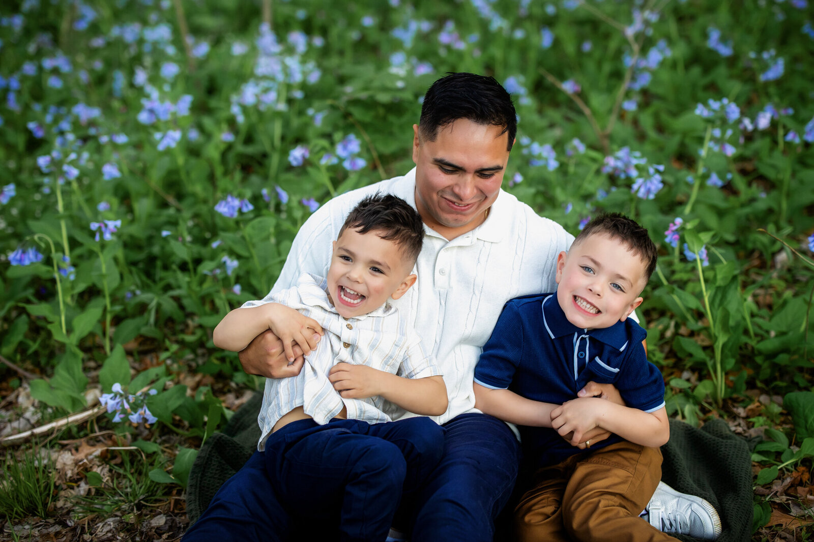 A man sits on a blanket outdoors surrounded by blue flowers, smiling with two young boys on his lap; all three appear happy and are laughing together in this charming outdoor portrait by a Bloomington family photographer.