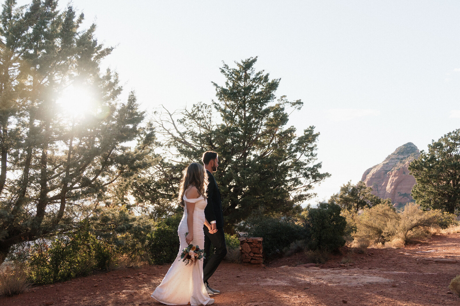 Bride and groom holding hands and walking through red dirt trails surrounded by pine trees during their elopement at Merry Go Round Rock in Sedona, Arizona, with sun flare and red rock formations in the background.