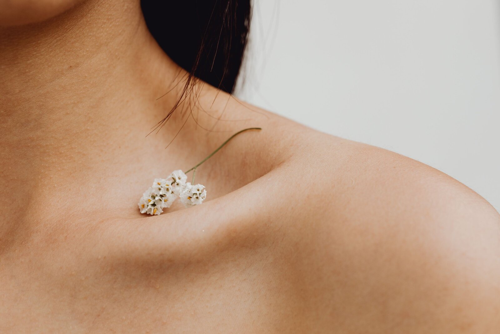 Close-up of a woman’s bare shoulder with a delicate white flower resting near her collarbone — symbolizing softness, self-care, and natural beauty