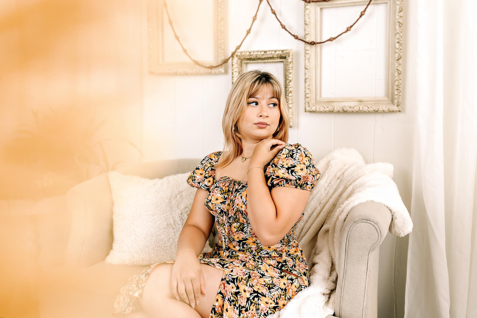 A person with blonde hair sits on a light-colored sofa, wearing a floral dress. They are gazing to the side, with an arm resting on the back of the sofa. The background features empty, ornate frames and gentle lighting.