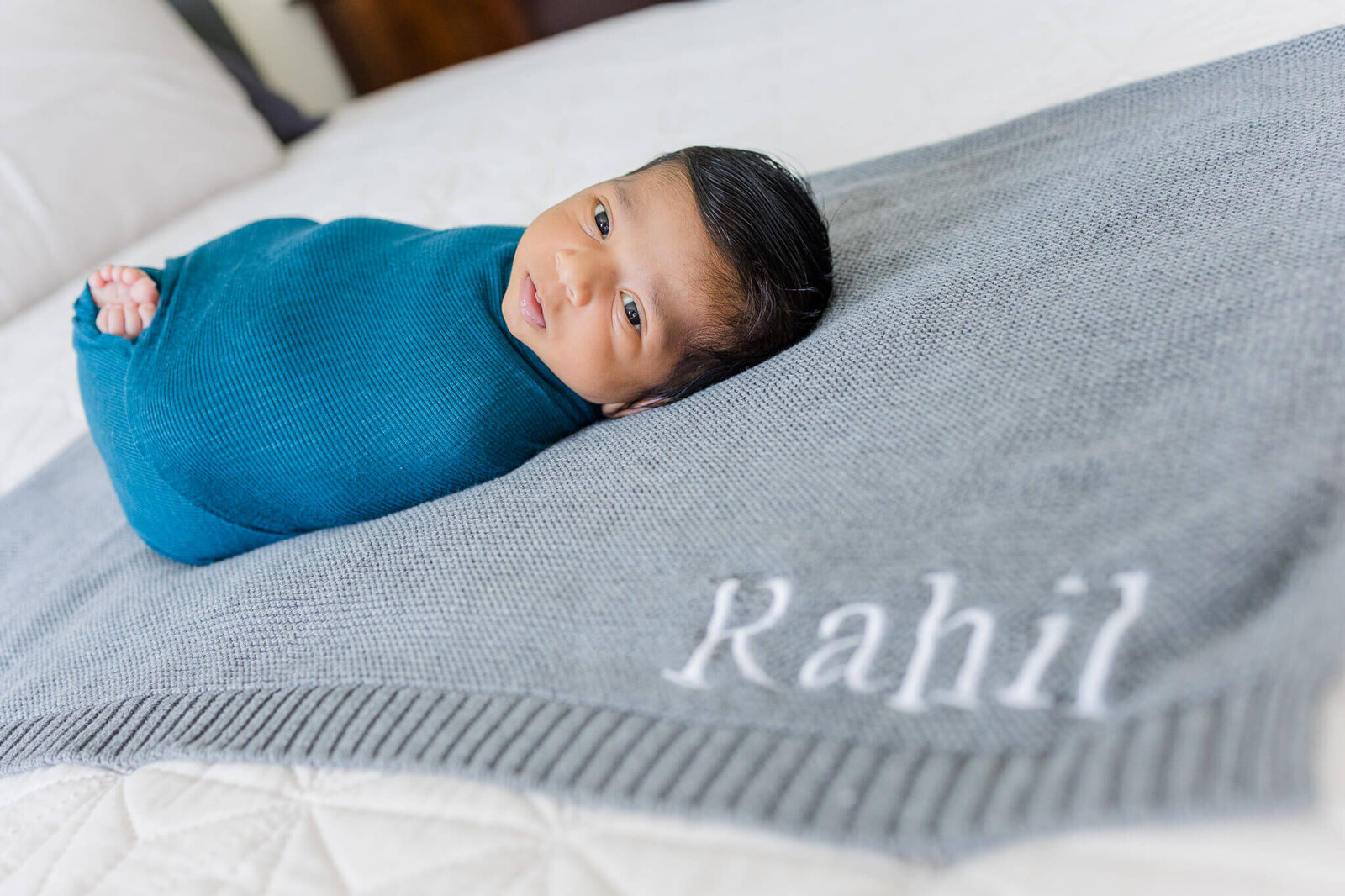A newborn boy wrapped in teal and laying on a gray blanket with is name on it.