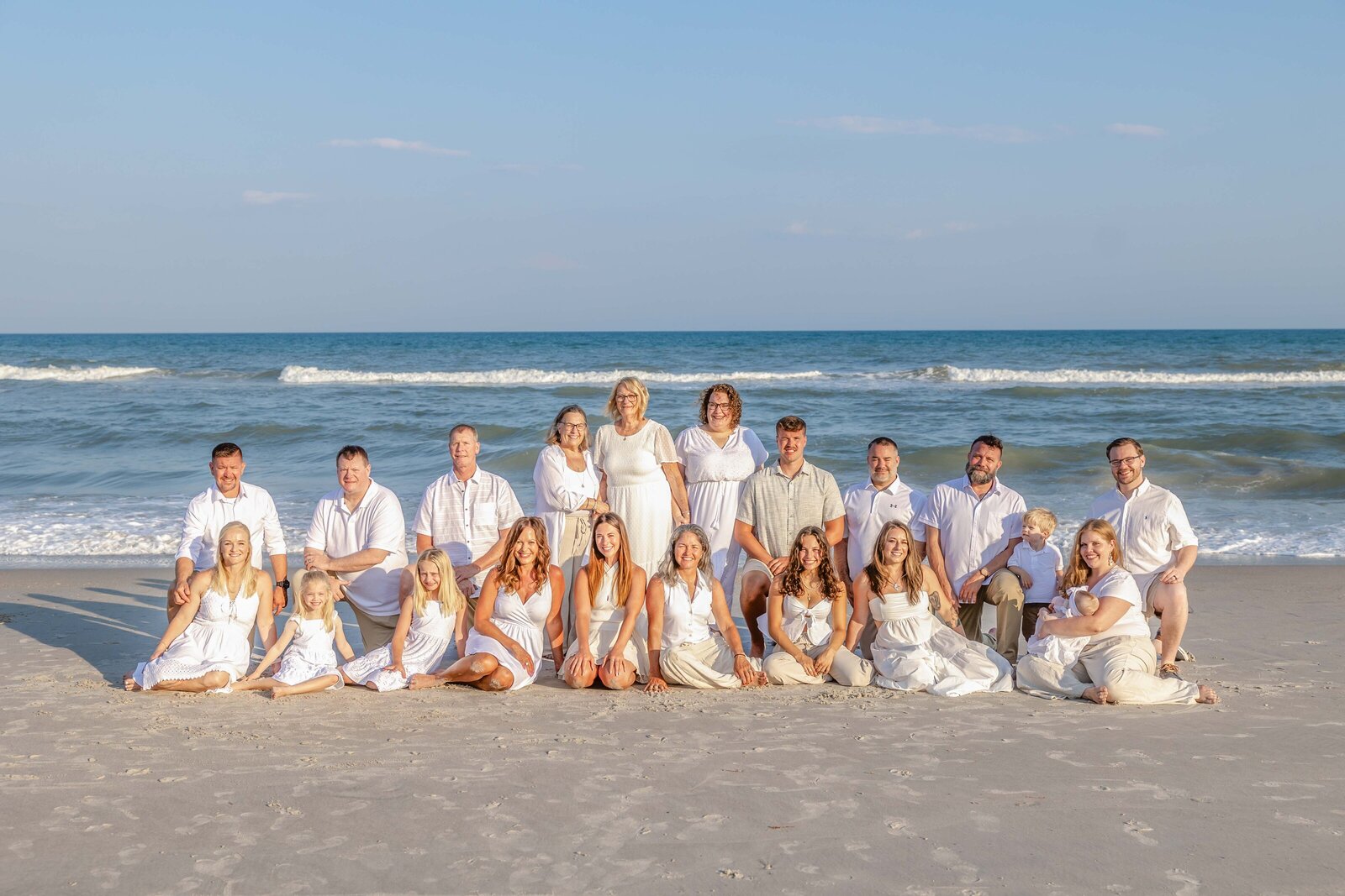 A large family dressed in white gathers on the sandy shores of Wrightsville Beach. The bright sunlight and ocean waves create a stunning backdrop, capturing the elegance of a family reunion. Ideal for destination family photographer NC, showcasing timeless beach gatherings.