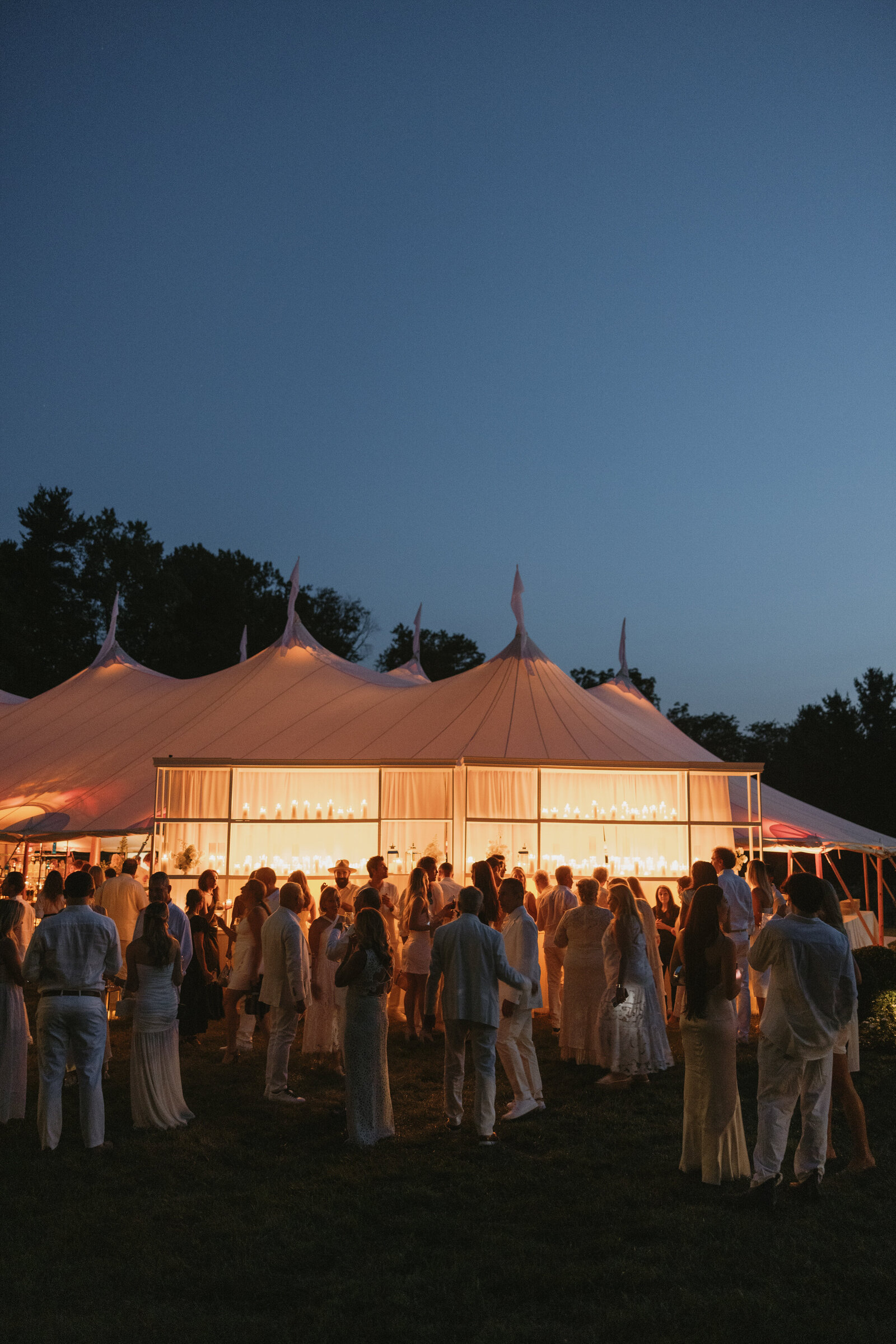 Large sailcloth wedding reception glowing at dusk with guests mingling in the foregroud
