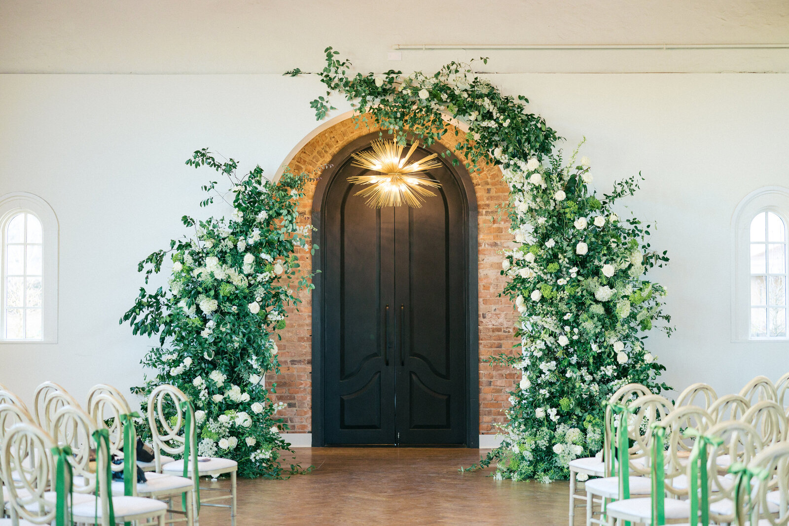 black arched door with gold sputnik chandelier exposed brick surrounding in the background, large green floral installation over the arch down to the ground