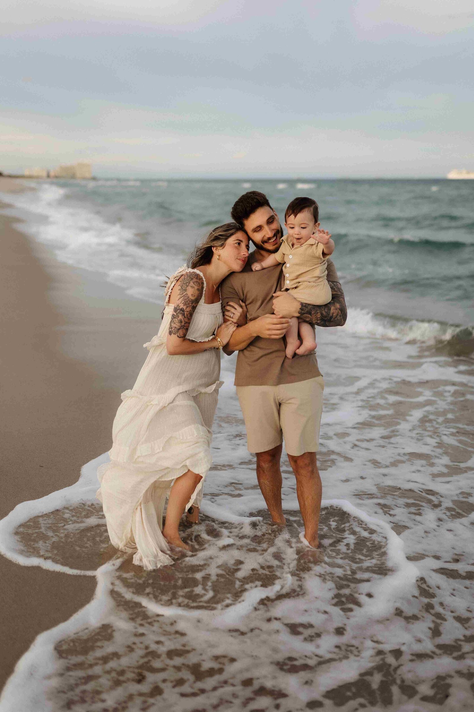 Miami family beach photo: parents and baby standing in gentle waves, ocean and sky in background