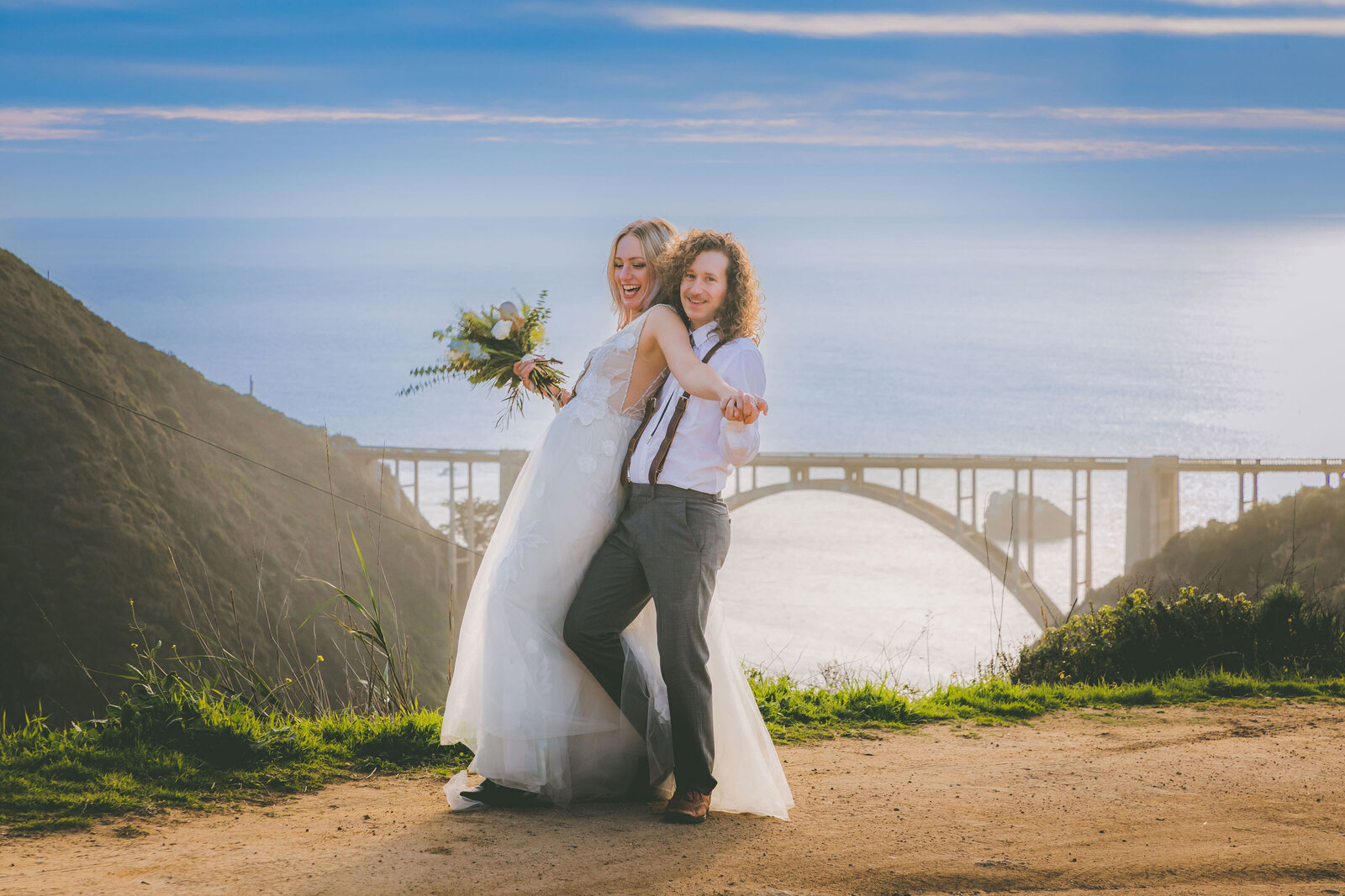 A wedded couple make silly poses with the Bixby Bridge in the background.