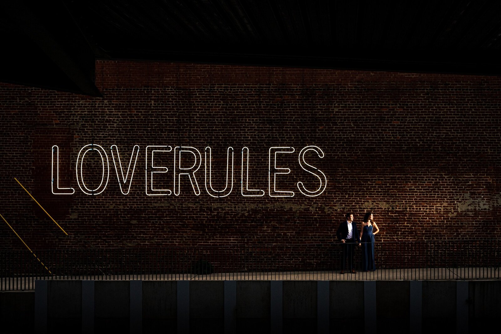 engaged couple poses in front of a neon sign that says Love Rules