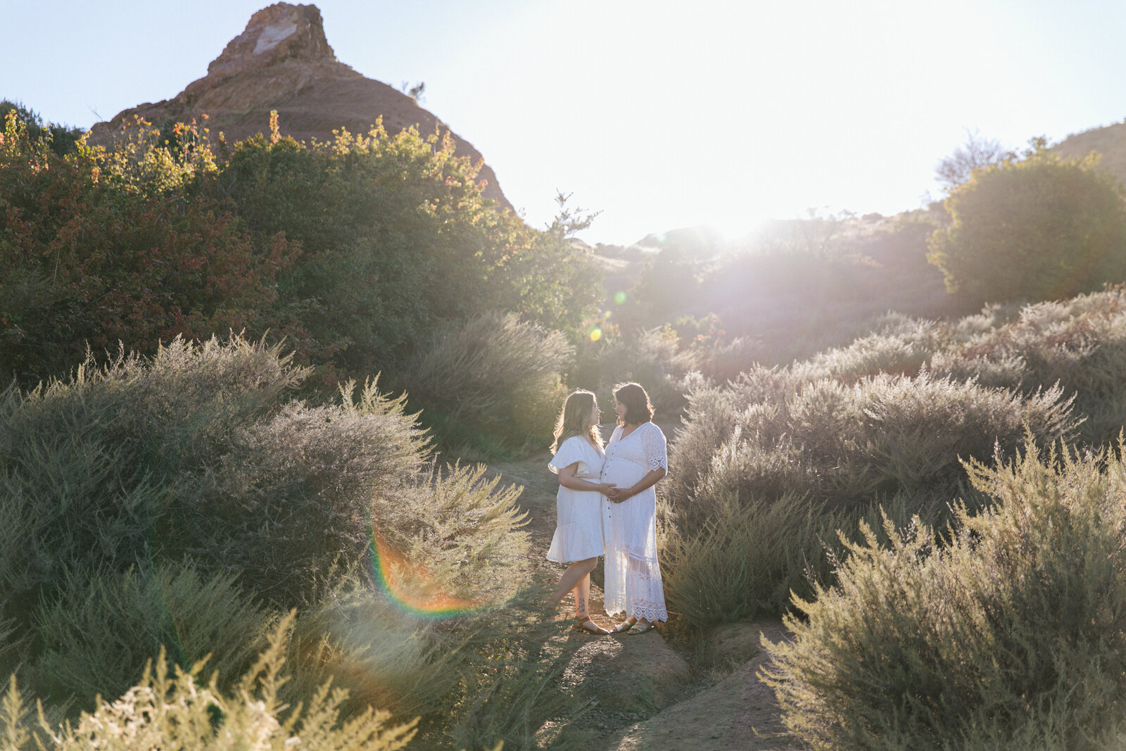 two moms wearing white, one of them pregnant, standing on a rocky hillside surrounded by bushes