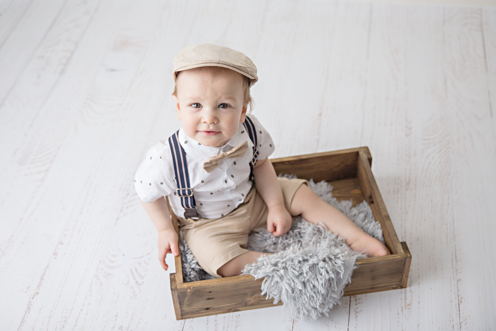 baby boy sitting in wooden crate with suspenders and hat on white wood floor