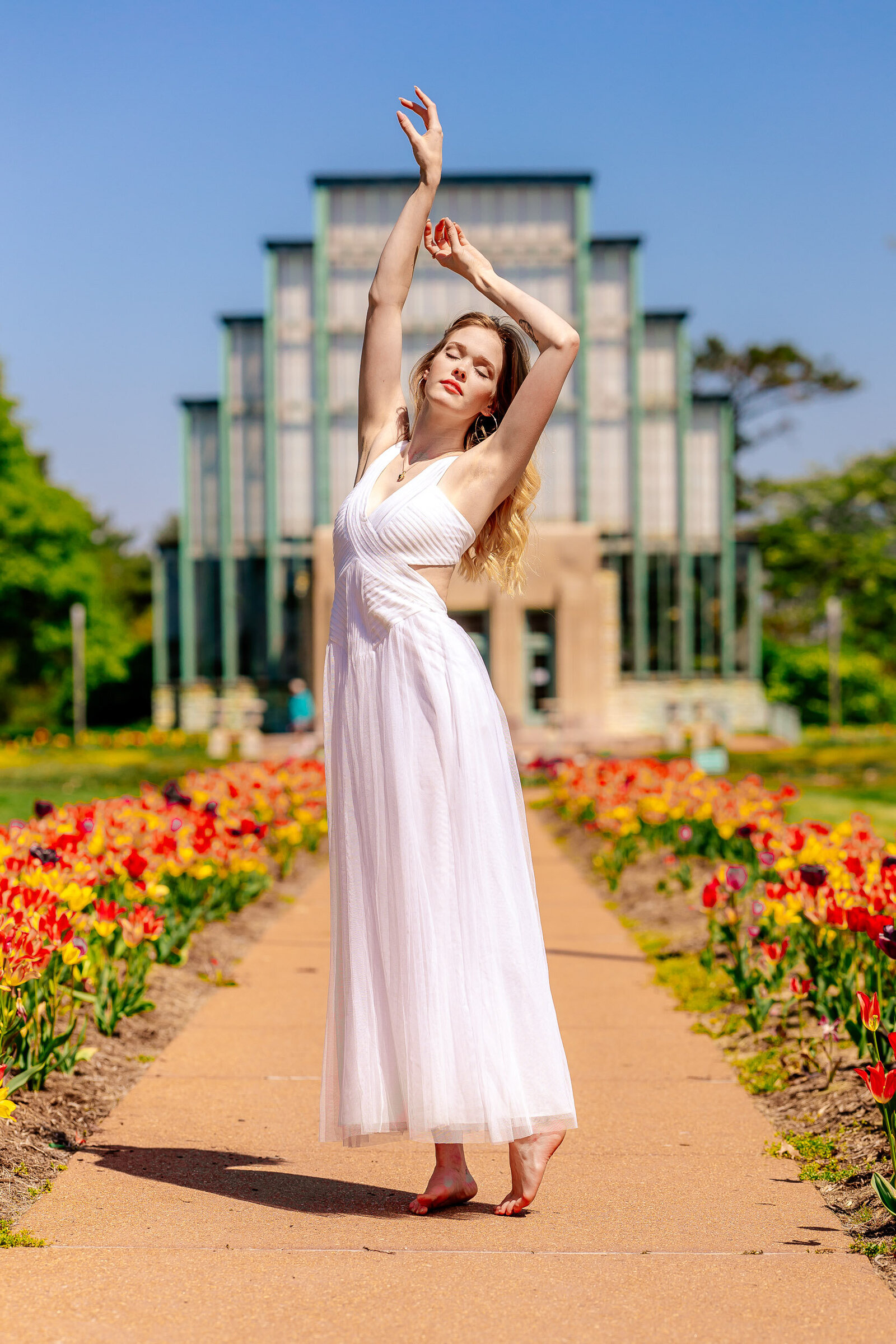 senior posing on tip toes in white dress with arms over head in front of the jewel box in forest park with tulips lining the path