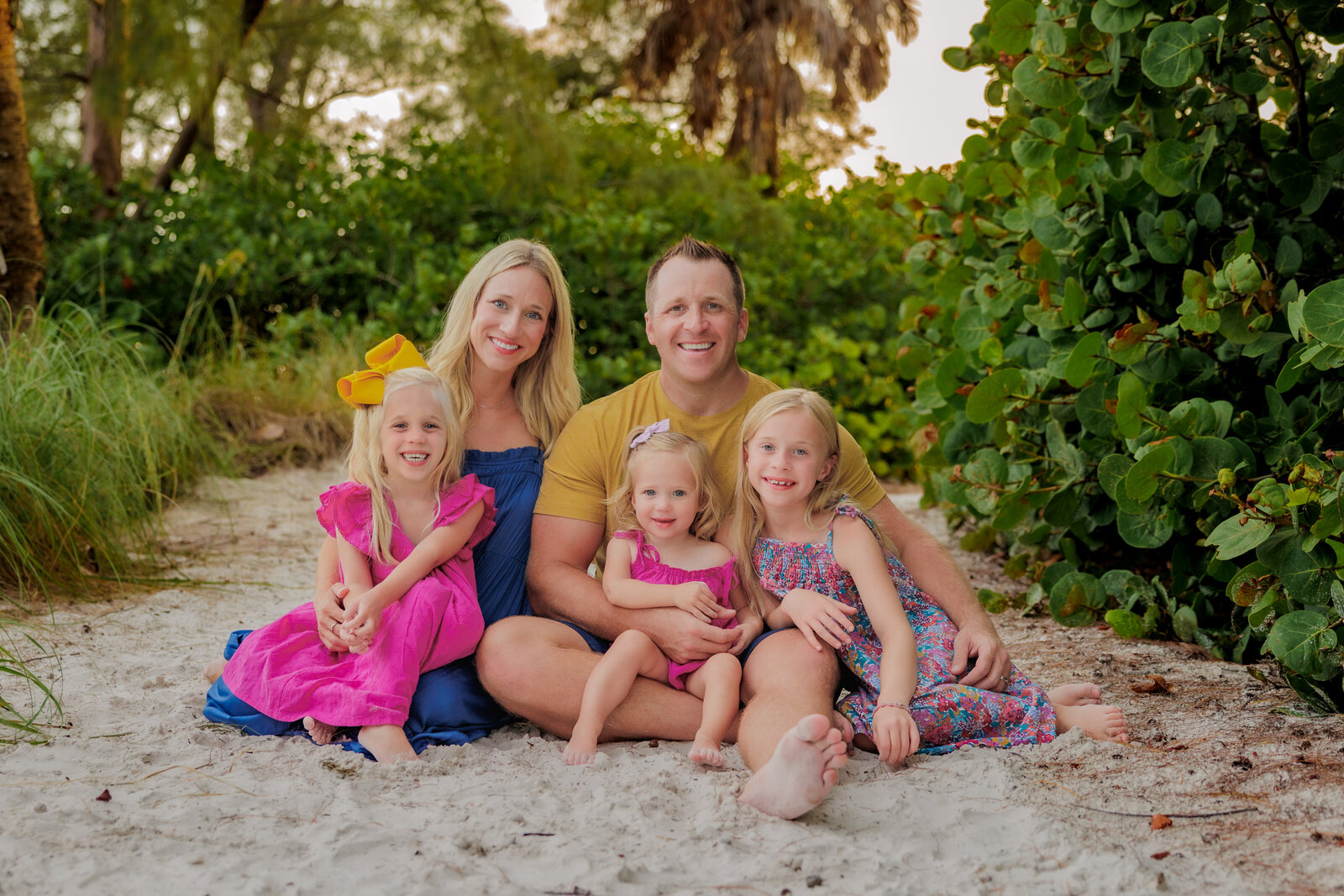 Family of five wearing a vibrant color scheme of blue, mustard yellow and fuchsia. They are sitting on a sand pathway with the children in the parents laps. They are surrounded by greenery and palm trees.