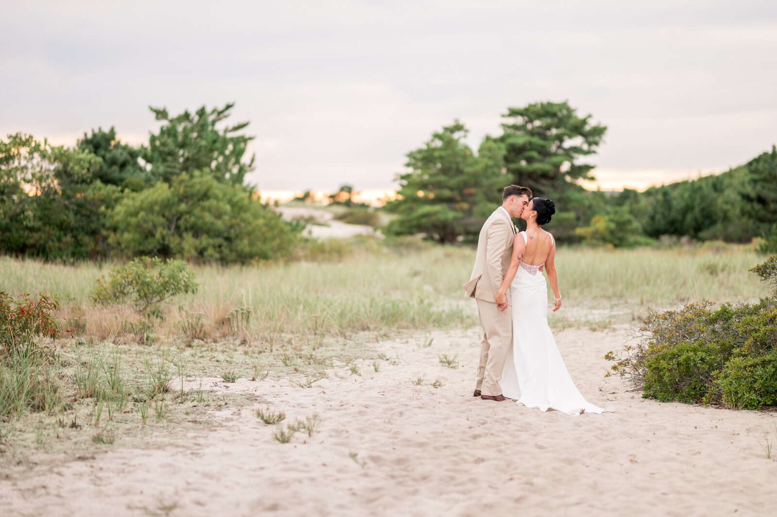 Bride and groom kissing on sandy beach path at Wychmere Beach Club surrounded by coastal greenery — soft, bright, airy New England wedding photography by Sarah Surette Photography