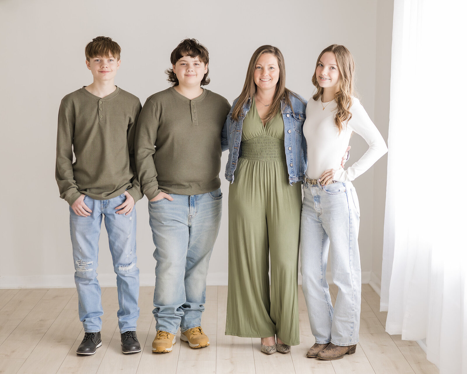 Family of 4 standing in a studio room on a light brown floor.  Two sons have dark brown hair, wearing dark green shirts, blue jeans, black shoes, and brown shoes, smiling at the camera.  Mom is wearing a blue jean jacket, has dark brown long hair, and a green jumpsuit with gray dress shoes, smiling at the camera.  Daughter has long brown hair, wearing a long sleeve white shirt, blue jeans, and dark brown shoes, smiling at the camera