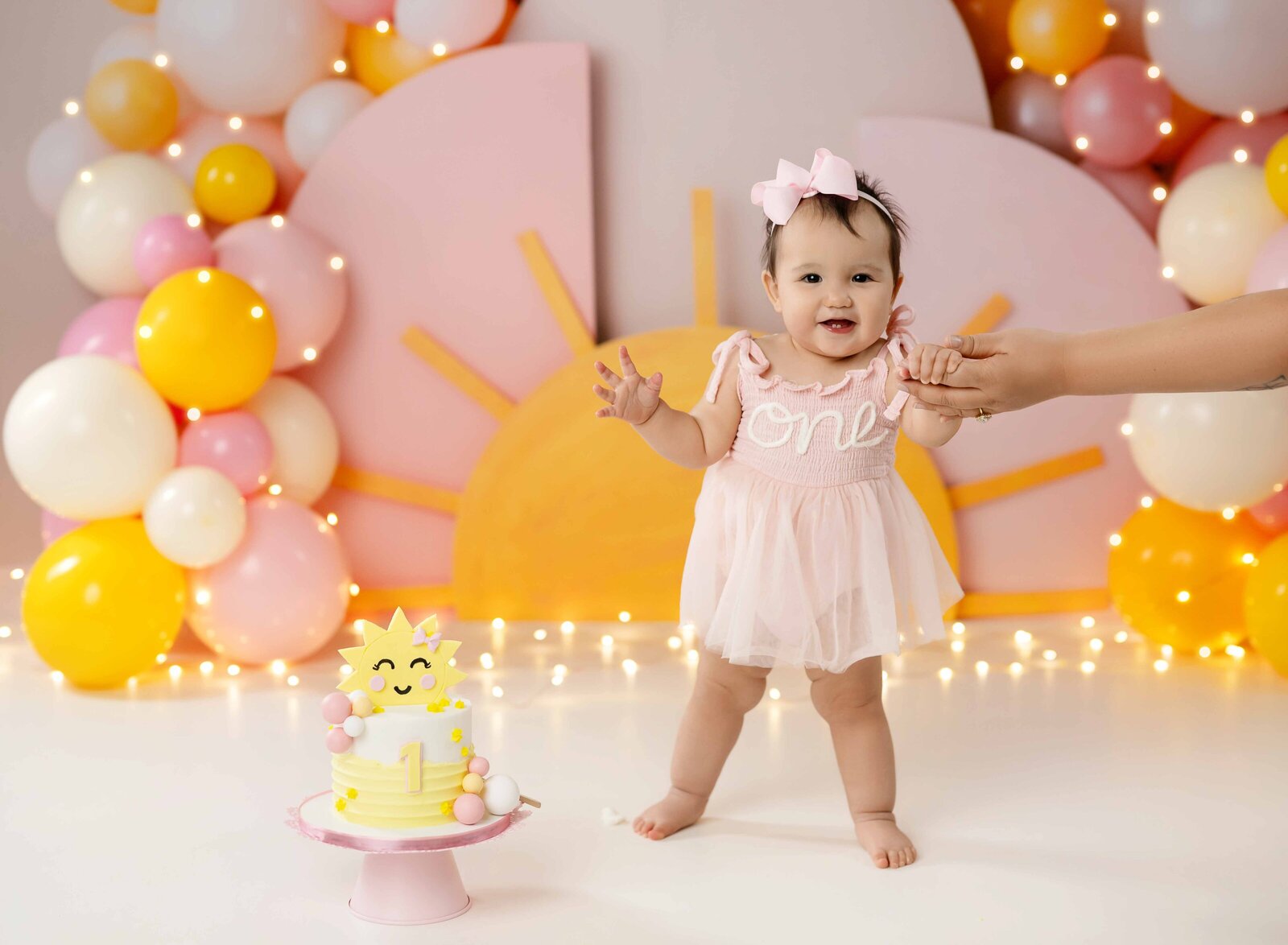 Baby taking cake smash photos and standing up, holding mom's hand, next to the pink and yellow sunshine cake.