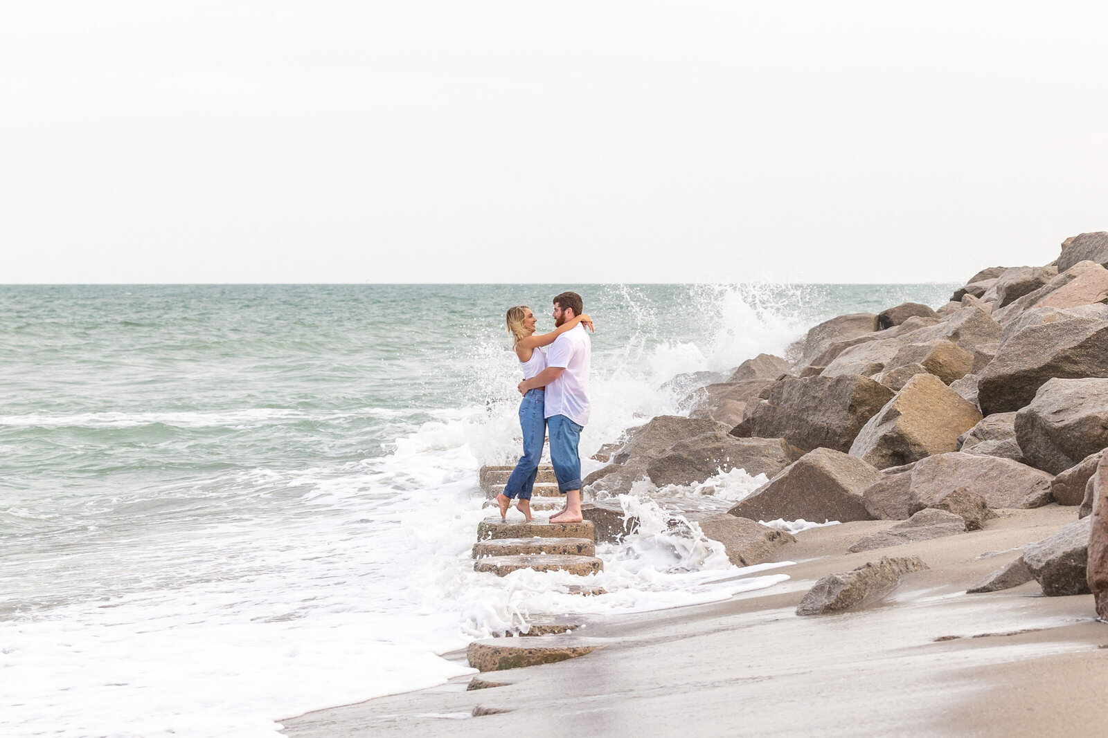 An engaged couple embracing on rocky steps by the ocean in Fort Fisher, NC, with waves crashing around them. This dynamic scene captures the thrill of authentic wedding photography, ideal for couples seeking unique and artistic engagement photos in North Carolina.