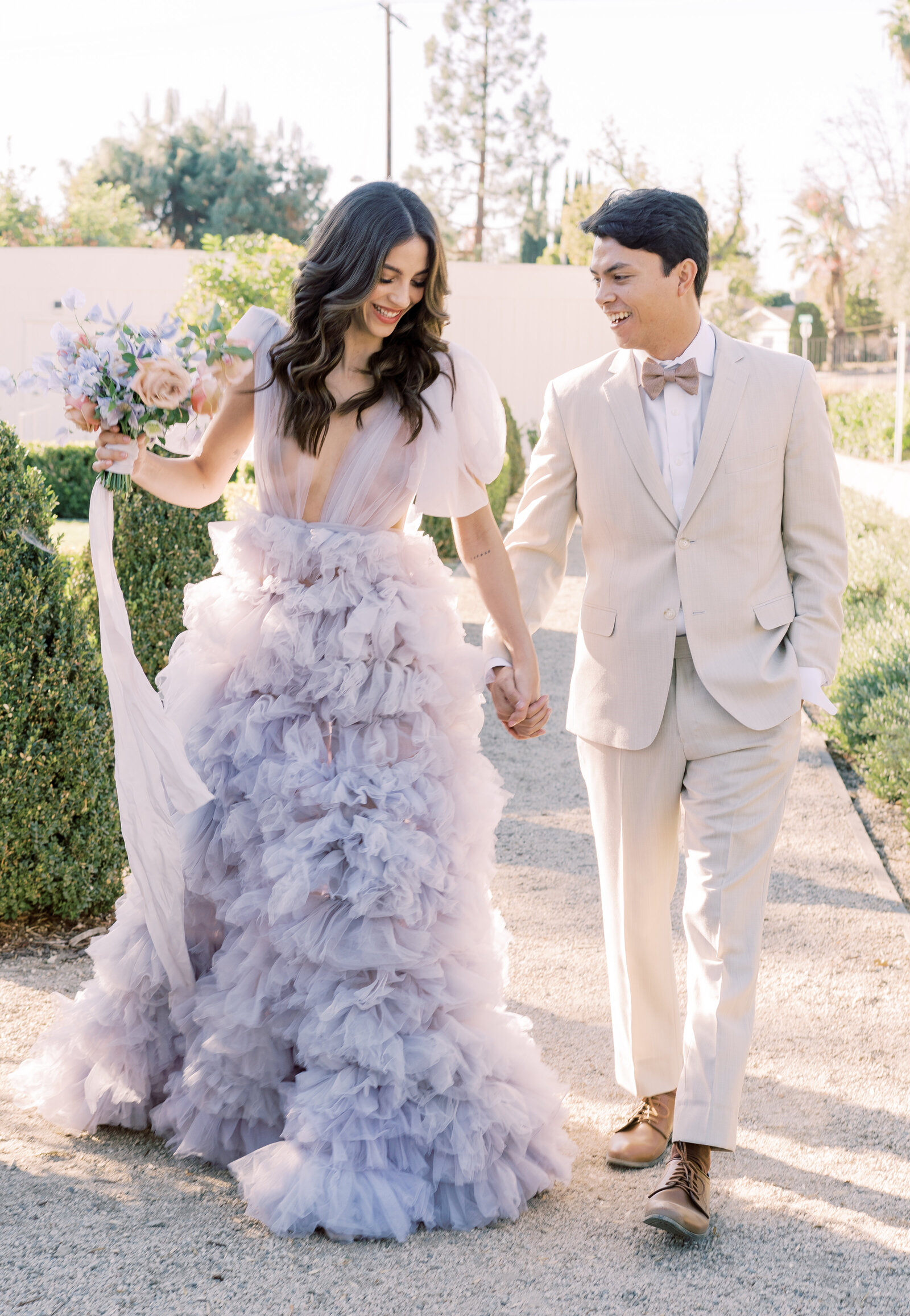 Portrait of bride and groom in a lavender wedding gown and cream suit walking in a garden outdoors with a bouquet in hand.