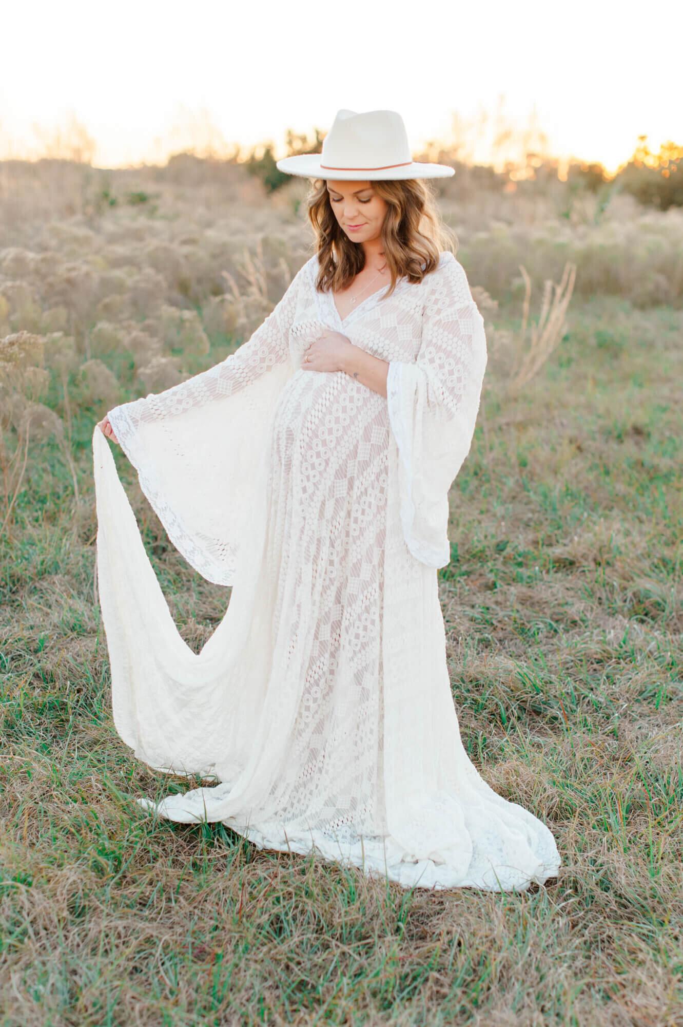 Pregnant mother holding her belly and standing in a white lace gown waving it in a tall grass field