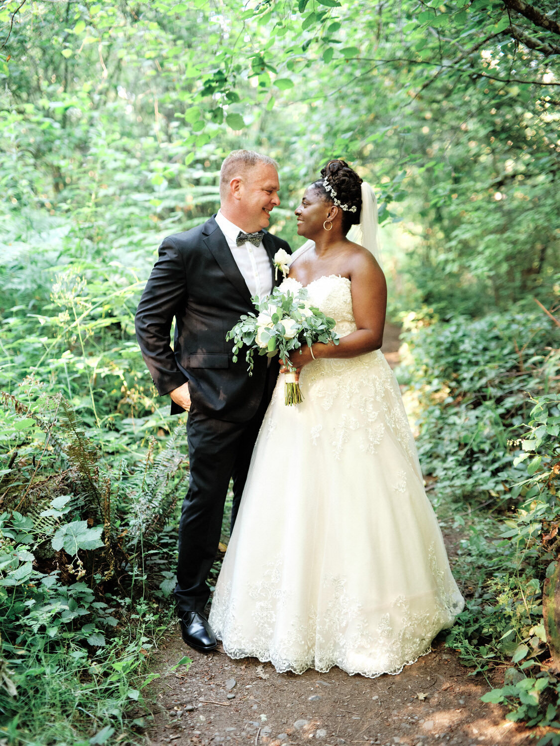 Bride and groom smile at each other in the woods surrounded by greenery