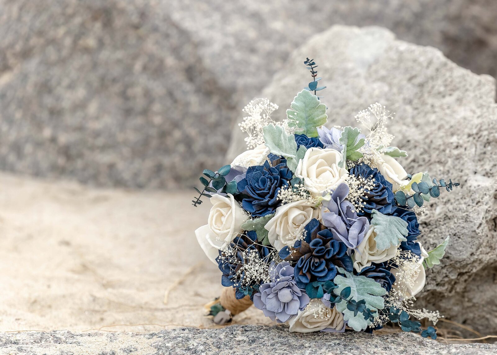 A blue and white wedding bouquet featuring roses and greenery rests on beachside rocks, captured during a Kure Beach, NC wedding, blending rustic coastal elements with elegant floral details.