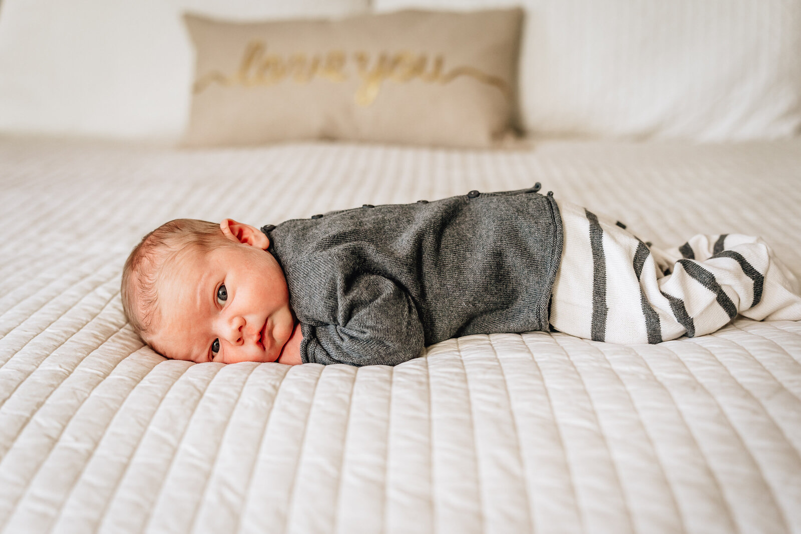 An adorable baby lies on a bed, facing the camera with curious eyes. The baby is dressed in a cozy outfit, with a pillow in the background that reads "love you" in cursive.