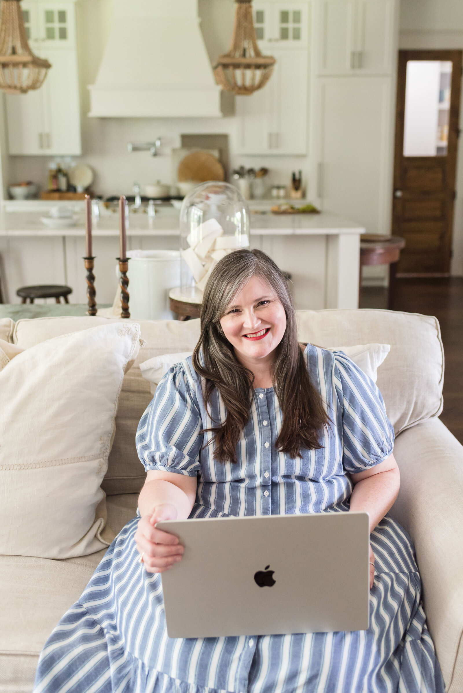 Lauren sits on her family room sofa holding her laptop in a blue and white striped dress