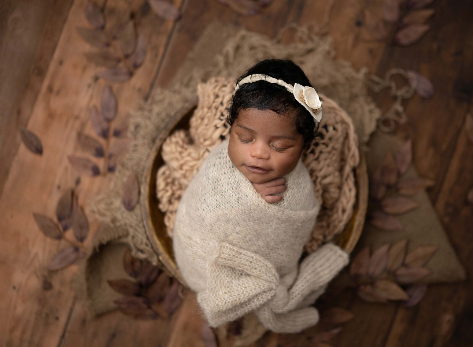 Newborn baby in cream swaddle, laying in a wooden bowl on wooden floor with fall leaves around bowl. Photographed by Jenni Nikki Photography at Marietta Newborn Photo Studio.