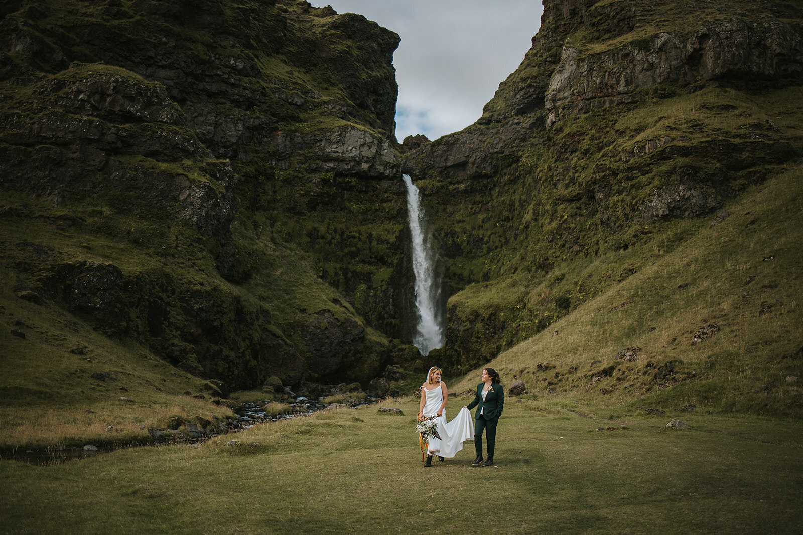 Couple on their elopement wedding day holding hands and walking with a waterfall behind them in Iceland.