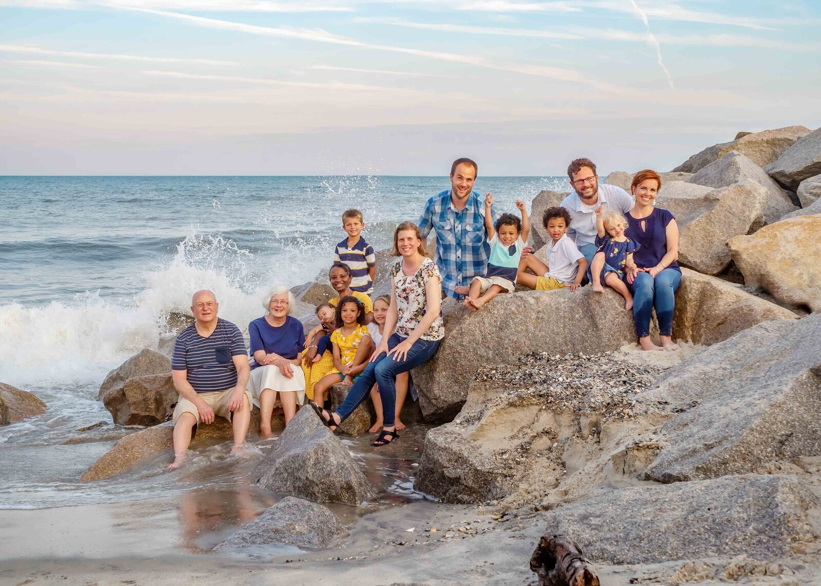 A large family sits on the rocky coastline at Fort Fisher, with ocean waves crashing behind them. Dressed in casual summer attire, they enjoy a lively and joyful moment. This image is perfect for destination family photographer NC, capturing dynamic family interactions by the sea.