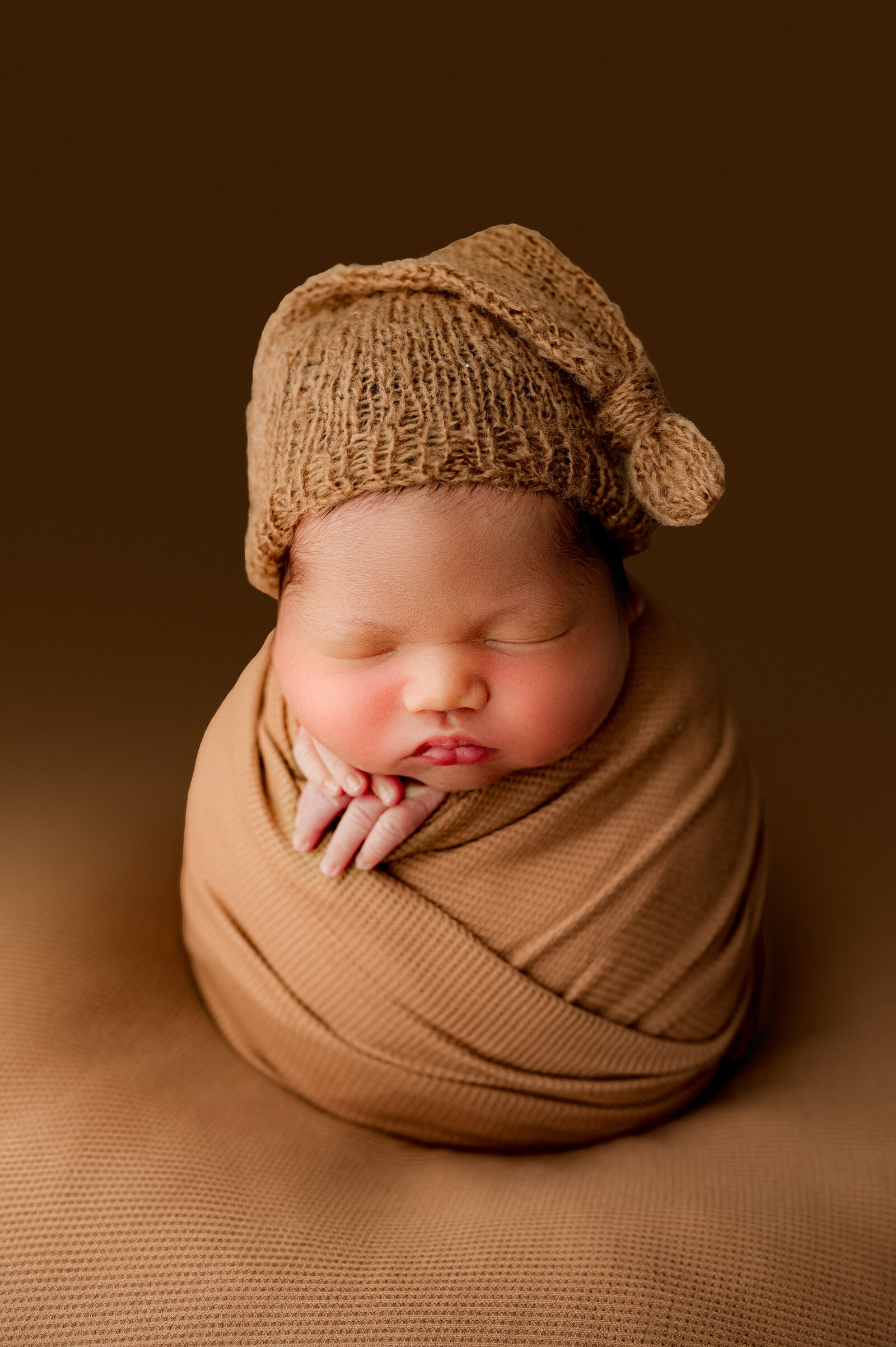 A newborn baby peacefully sleeping, wrapped in a light brown blanket. The baby wears a knitted brown hat with a small decoration on top. The warm brown background creates a cozy and unified look, perfectly captured by Charlottes newborn photographer in their studio.
