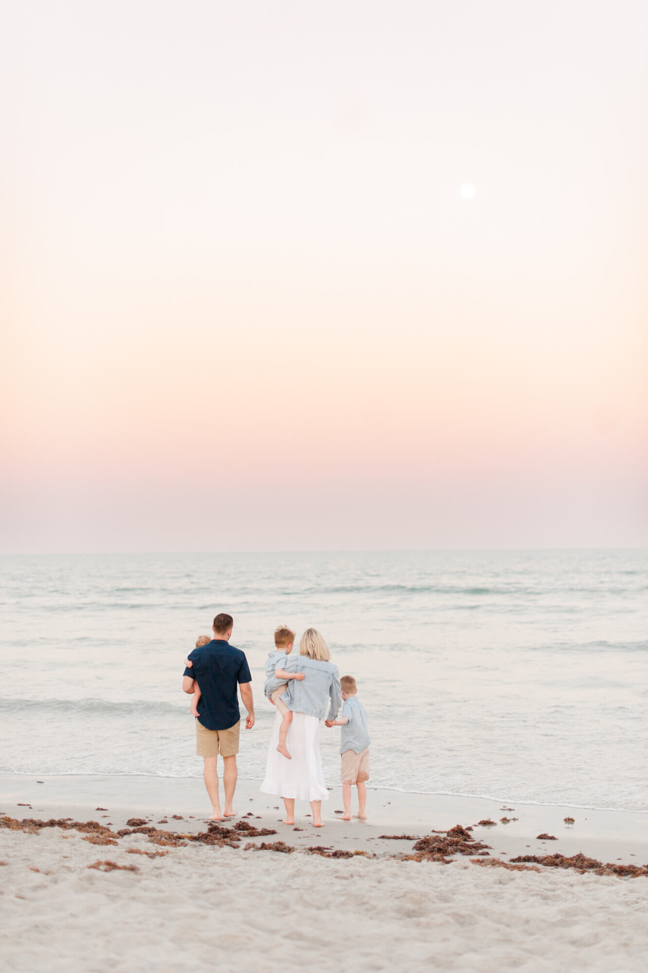 Family stands near the shoreline looking at the moon and watching the waves crash.