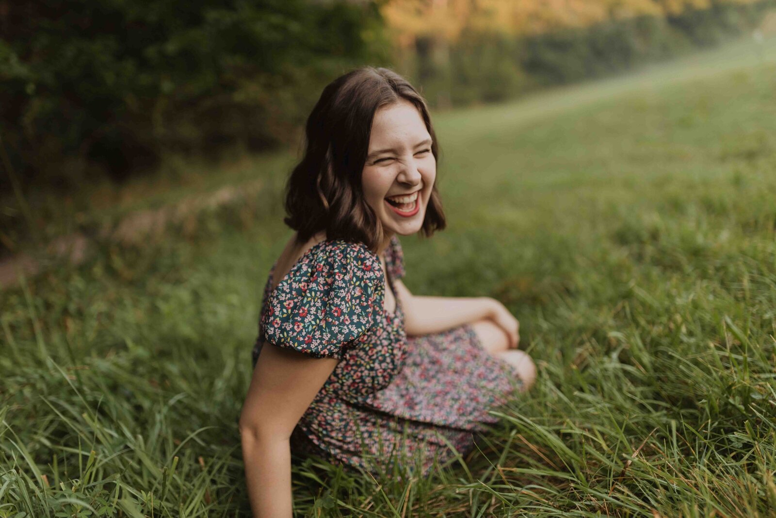 Senior girl is wearing a sun dress, sitting in grass. Back is to the camera and she looks back laughing with eyes closed.