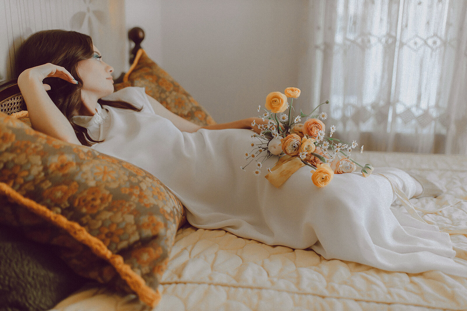 Editorial bridal portrait of a modern bride in a vintage-inspired silk gown, reclining on a bed with a warm-toned bouquet and cinematic styling.