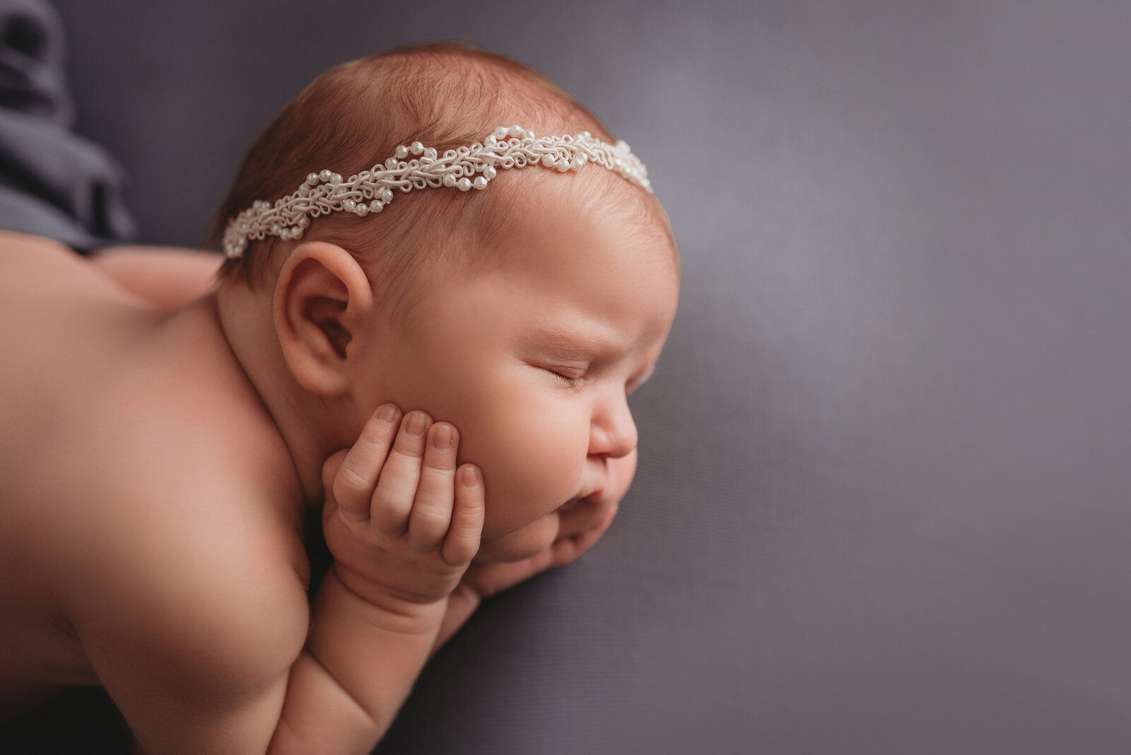 Newborn girl nude laying on side wearing a pearl headband with hands on cheeks on blue fabric backdrop