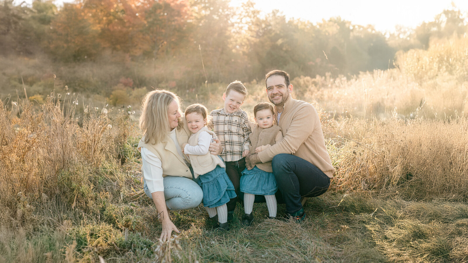 A candid family portrait captured in the soft, golden morning light of an open field. The image features a mother, father, and their three young children, including twin girls in denim skirts and cozy vests, all smiling and embracing each other. The gentle sunlight enhances the natural and joyful atmosphere, embodying the warm and emotive style of KCP Families' photography.