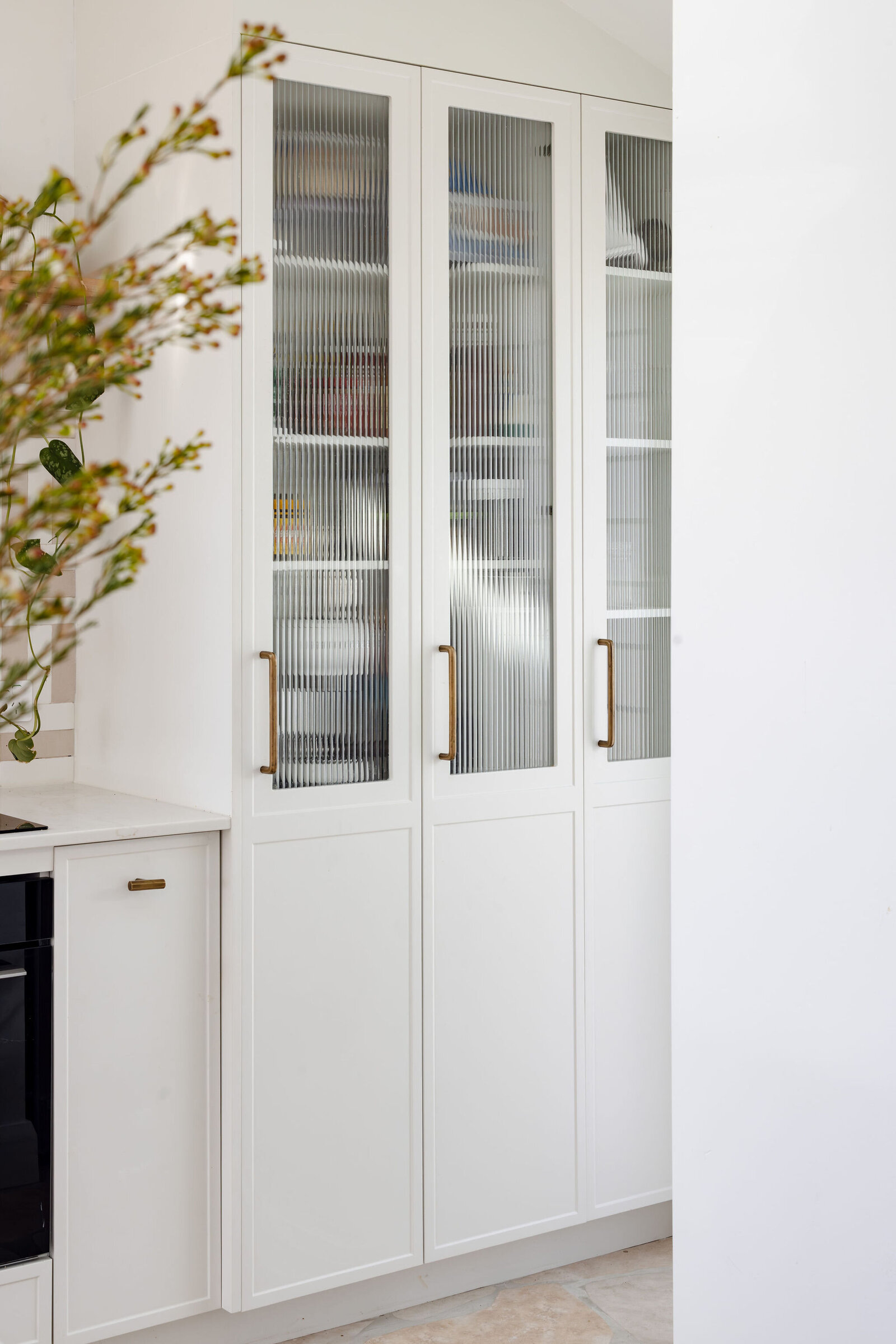 White kitchen cabinets with glass doors and brass handles exude elegance, enhanced by a plant on the left. Discover this exquisite touch in Newcastle Kitchen and Bathroom Designer collections, where style meets functionality.