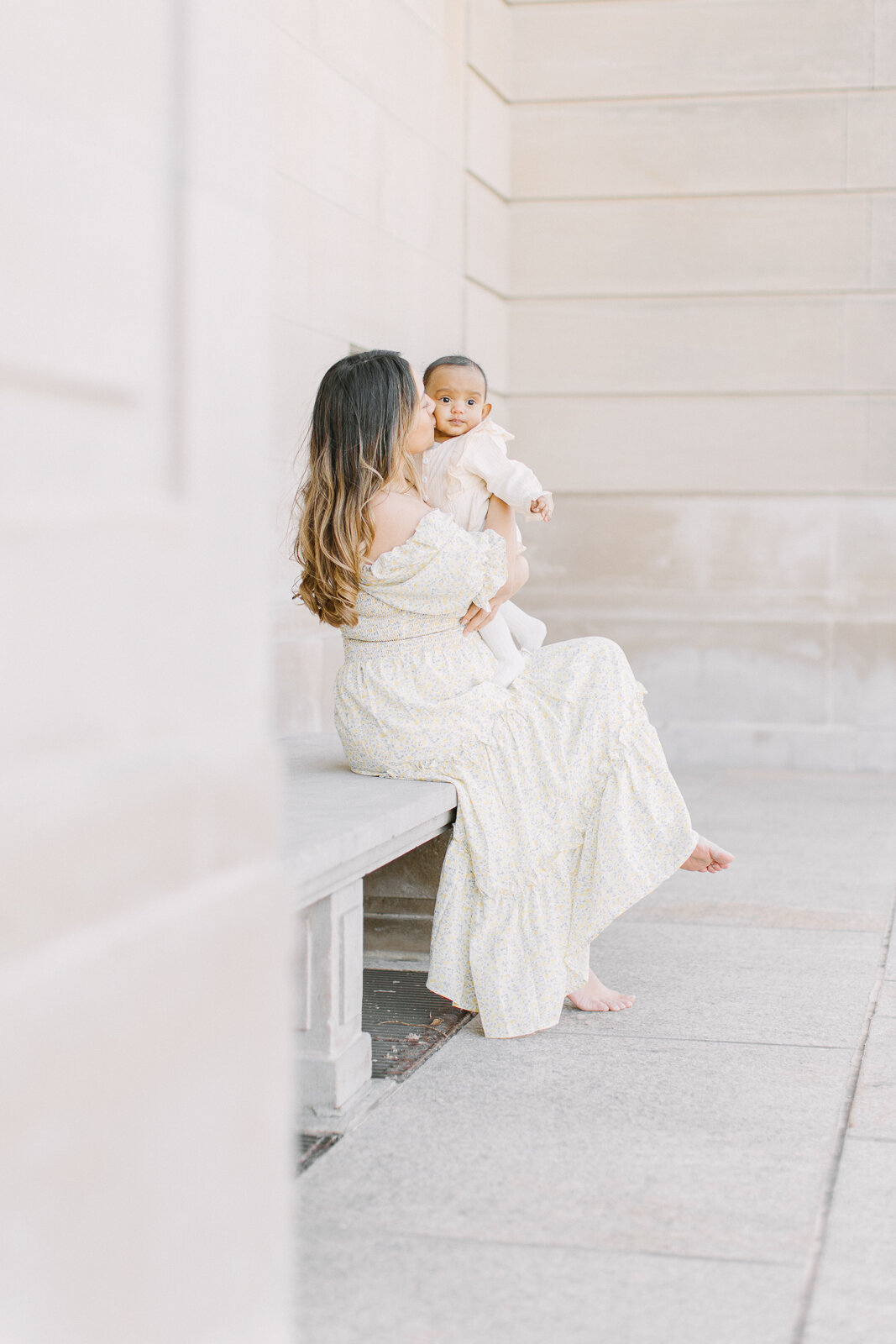 A young mother holding her baby girl while sitting on a bench at the Arlington Town Hall during photo session with Boston photographer Corinne Isabelle