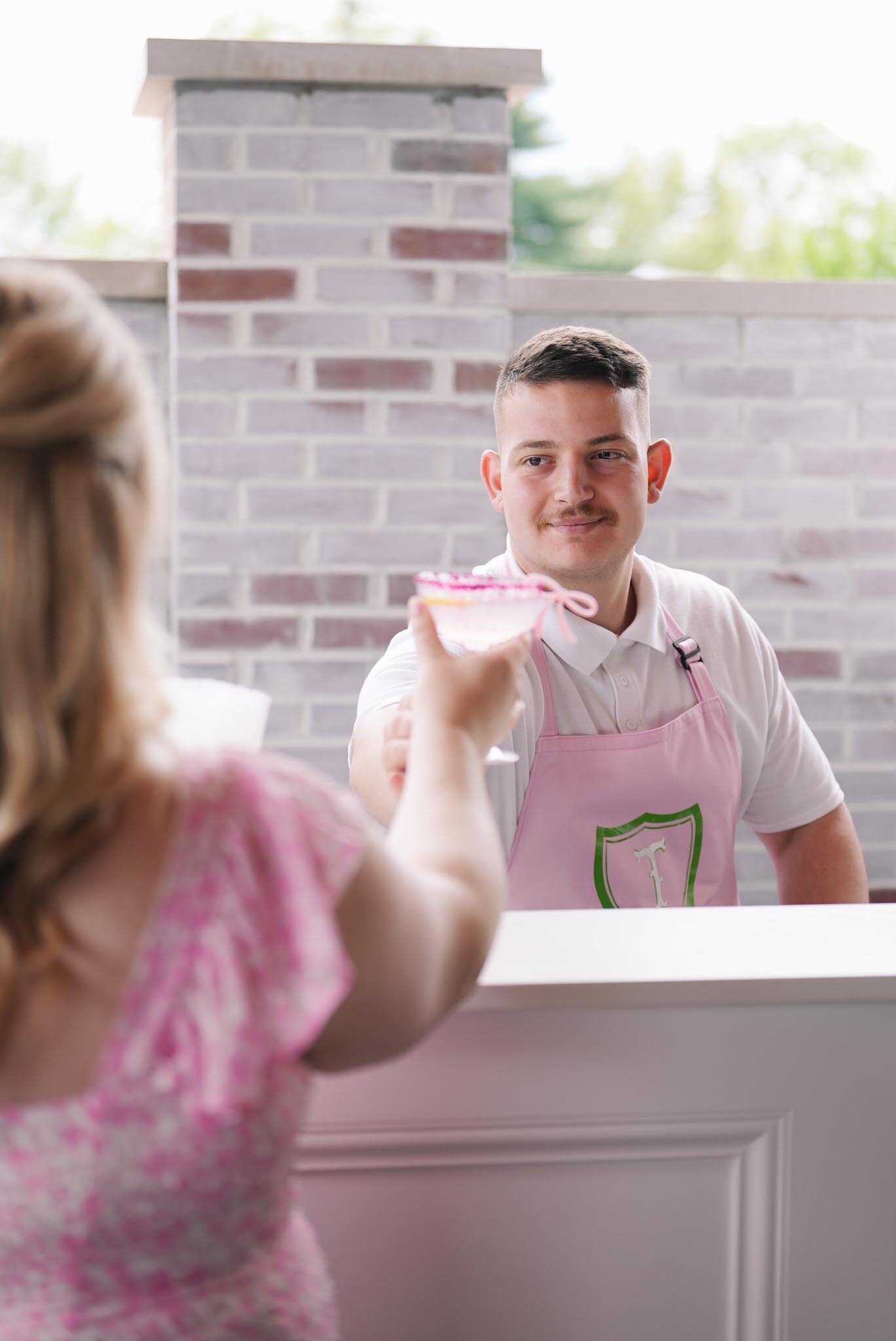 A man wearing a pink apron hands a pink cocktail to a woman