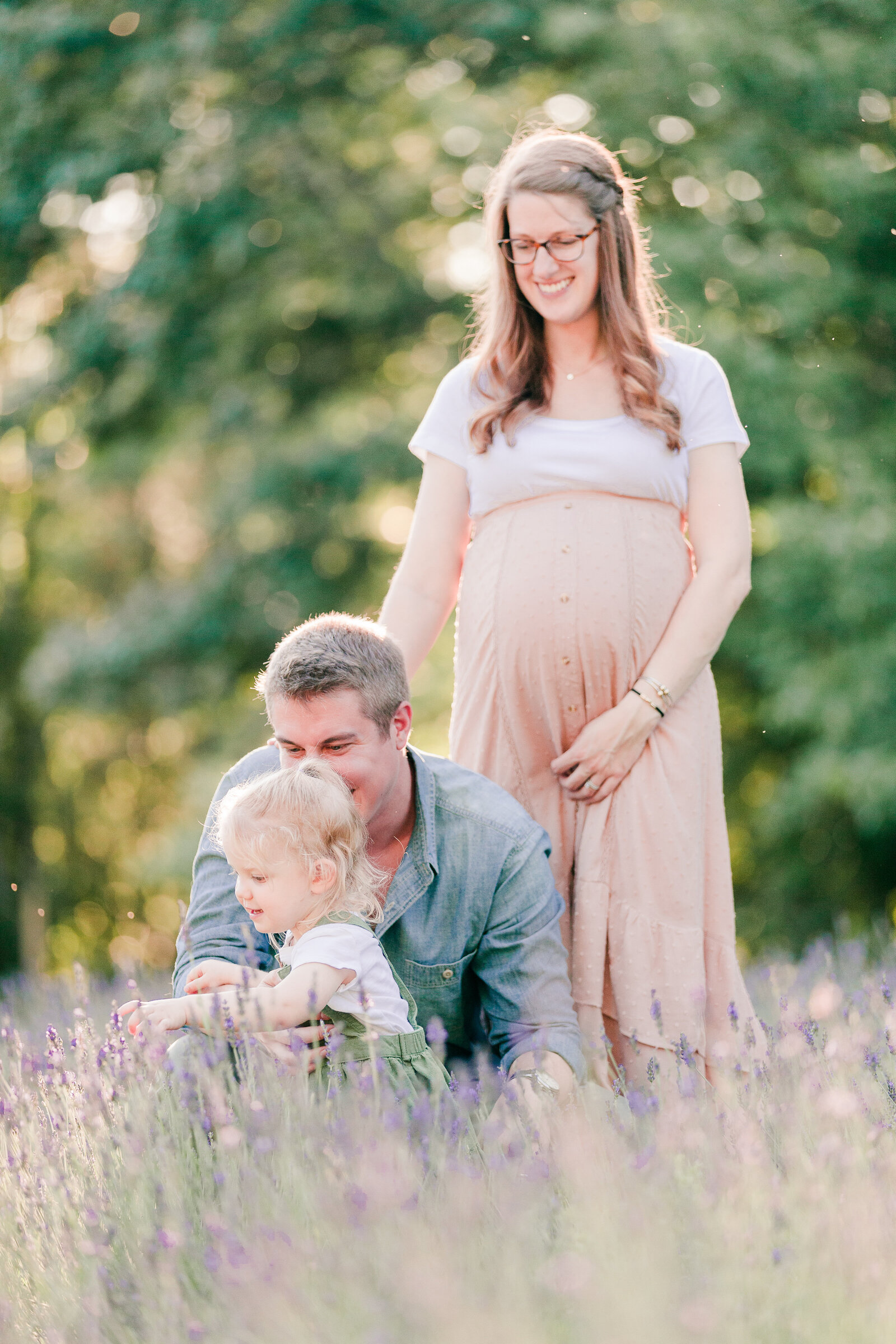 Pregnant mom standing and holding her baby bump, smiling down at her husband and three year old kneeling in a lavender field by NH newborn photographer Fieldstone Studio.