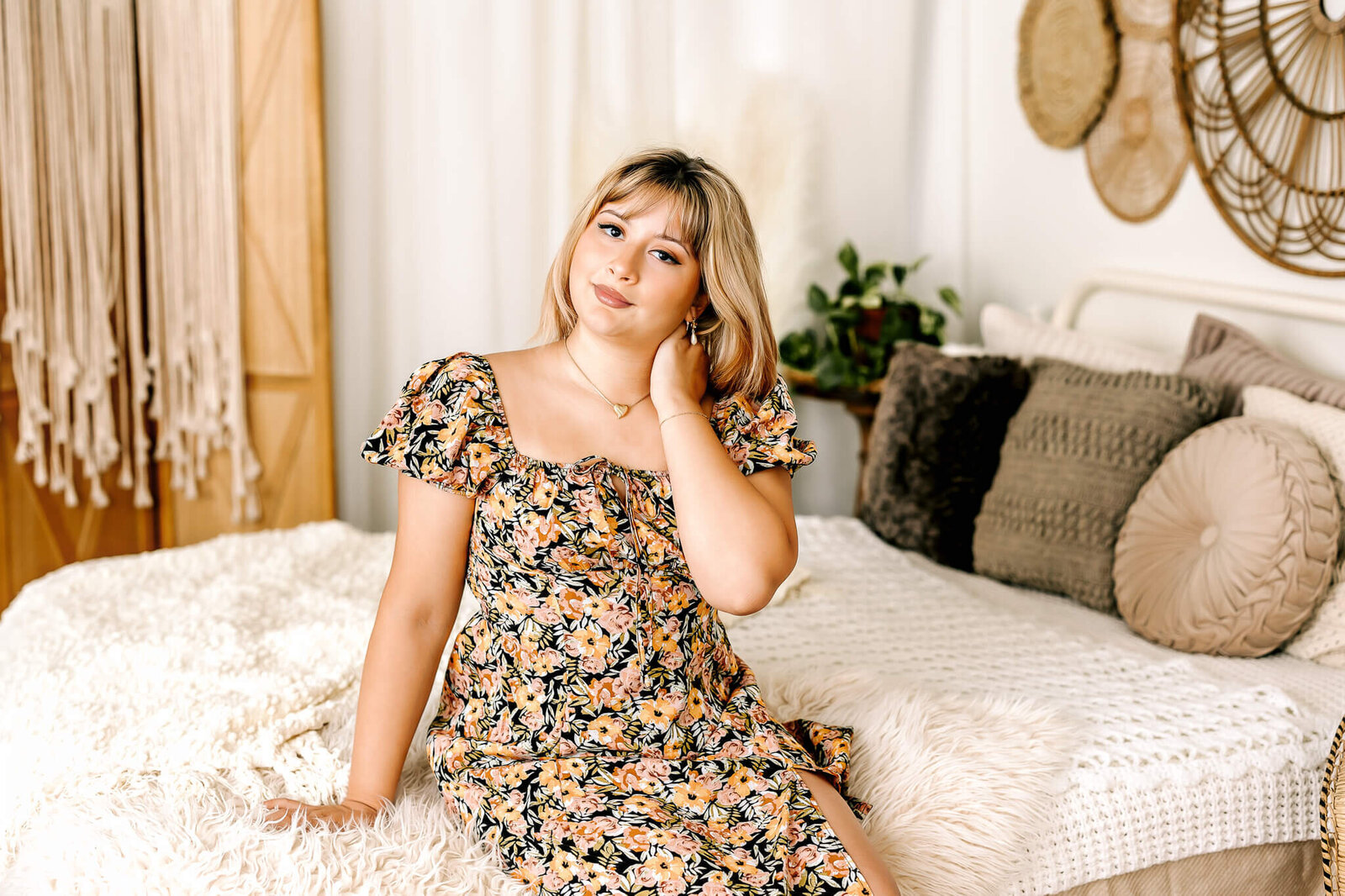 A person in a floral dress sits on a cozy bed with textured pillows and a fur throw, surrounded by bohemian decor including a macrame wall hanging and woven baskets. They have a relaxed and thoughtful expression.
