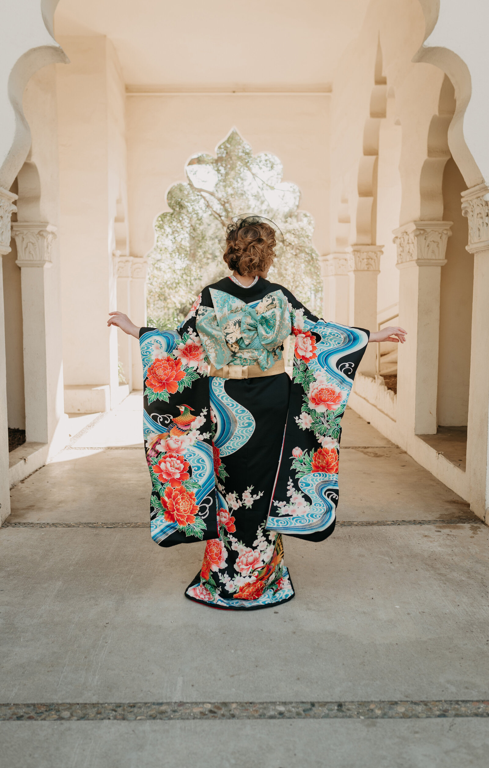 Back view of bride wearing elegant black hikifurisode wedding kimono rental with flowing peony and pheasant motif, posing under the Moorish arch at Brand Park Glendale, Los Angeles California | ブランドパーク グレンデール 黒引き振袖レンタル 後ろ姿 豪華牡丹と雉柄