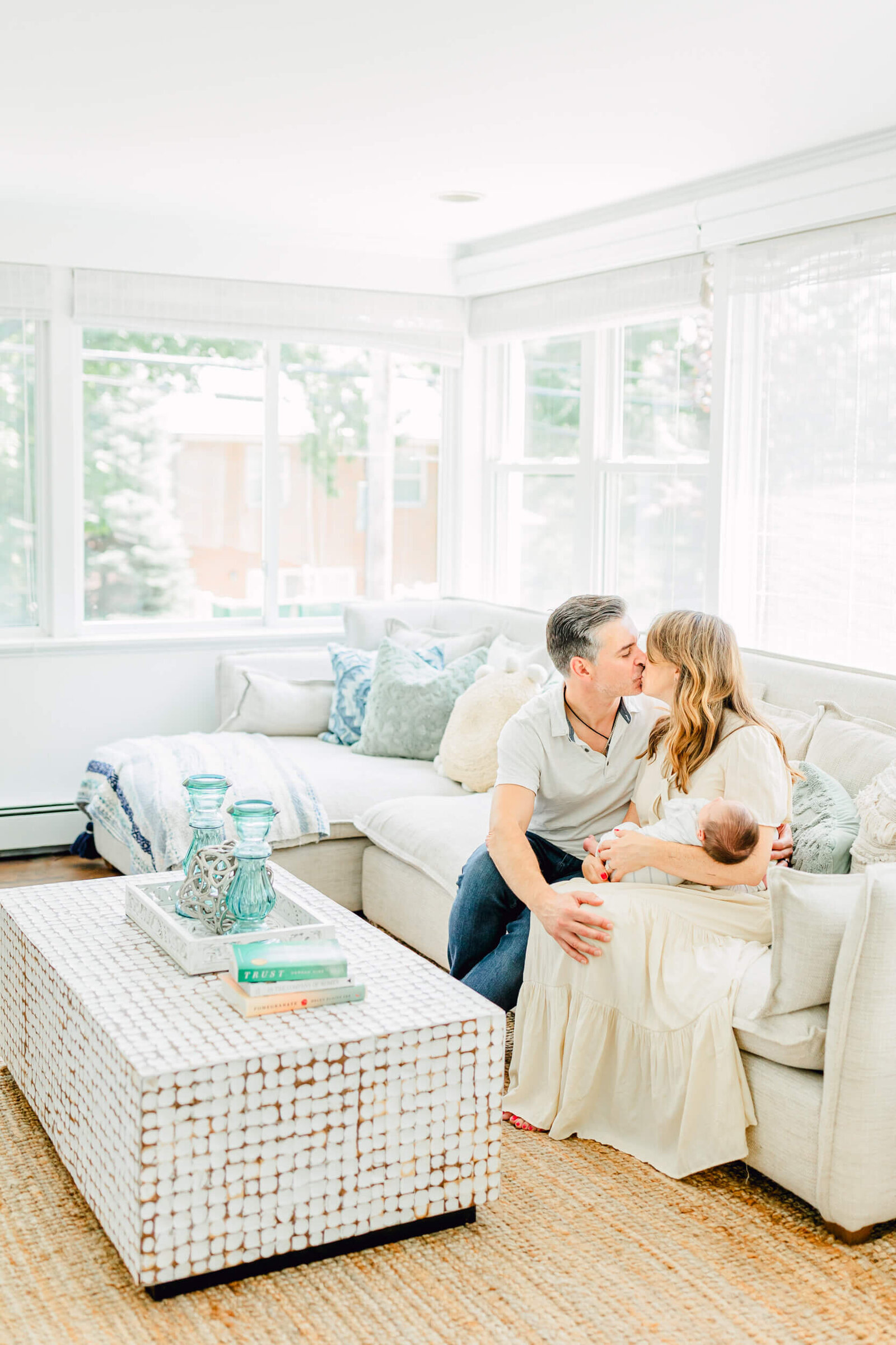 Mom and dad share a kiss on the couch while mom holds a newborn