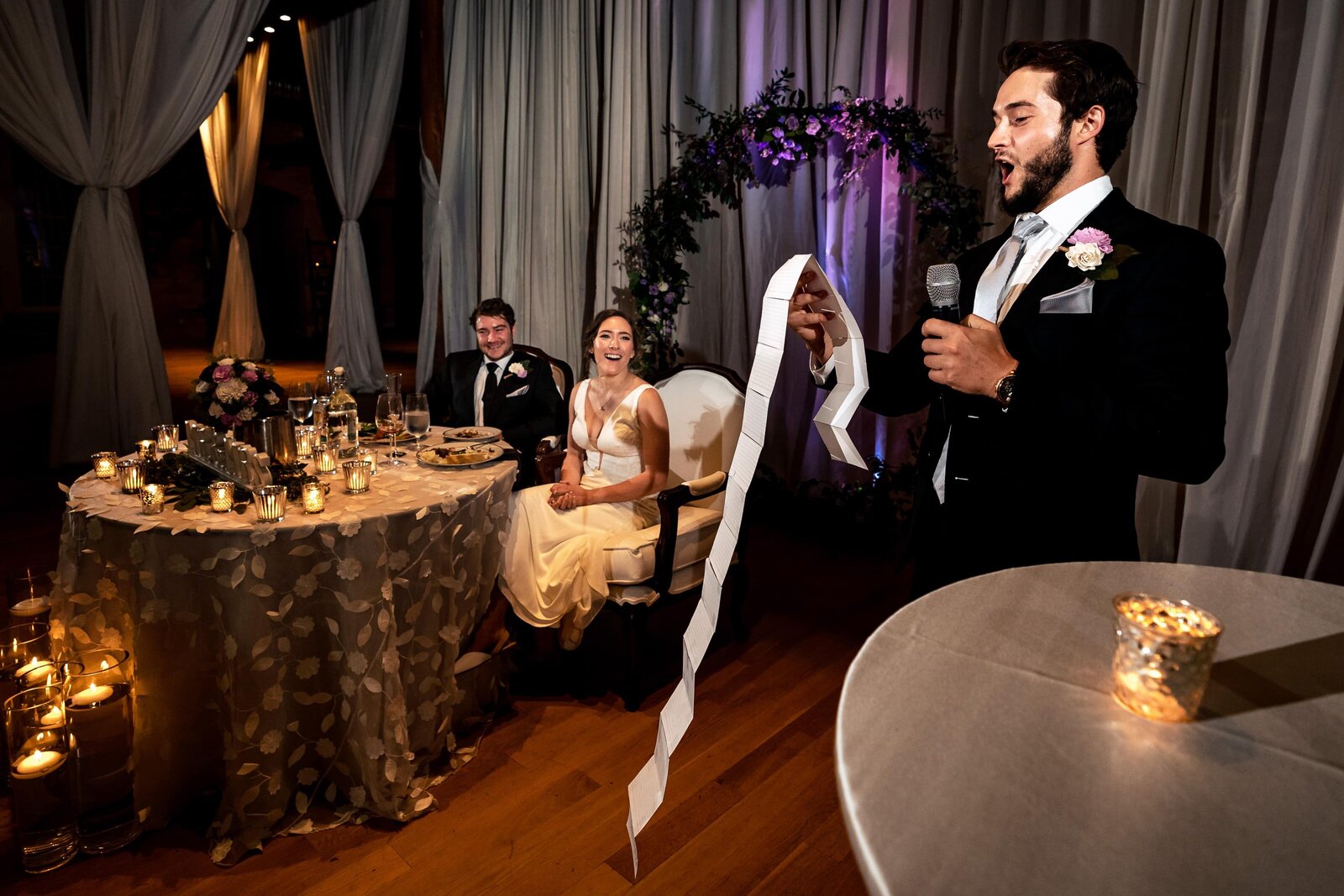 wedding couple sitting at their sweetheart table laughs as a groomsman makes a toast in the foreground