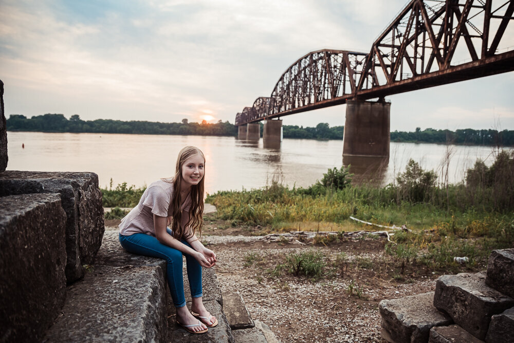 henderson train bridge and river front behind senior sitting in pink shirt smiling by klem photography senior photographer in henderson ky