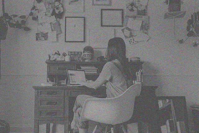 A web designer sits at a desk with a laptop in a room adorned with various papers, frames, and lush plants on the walls.